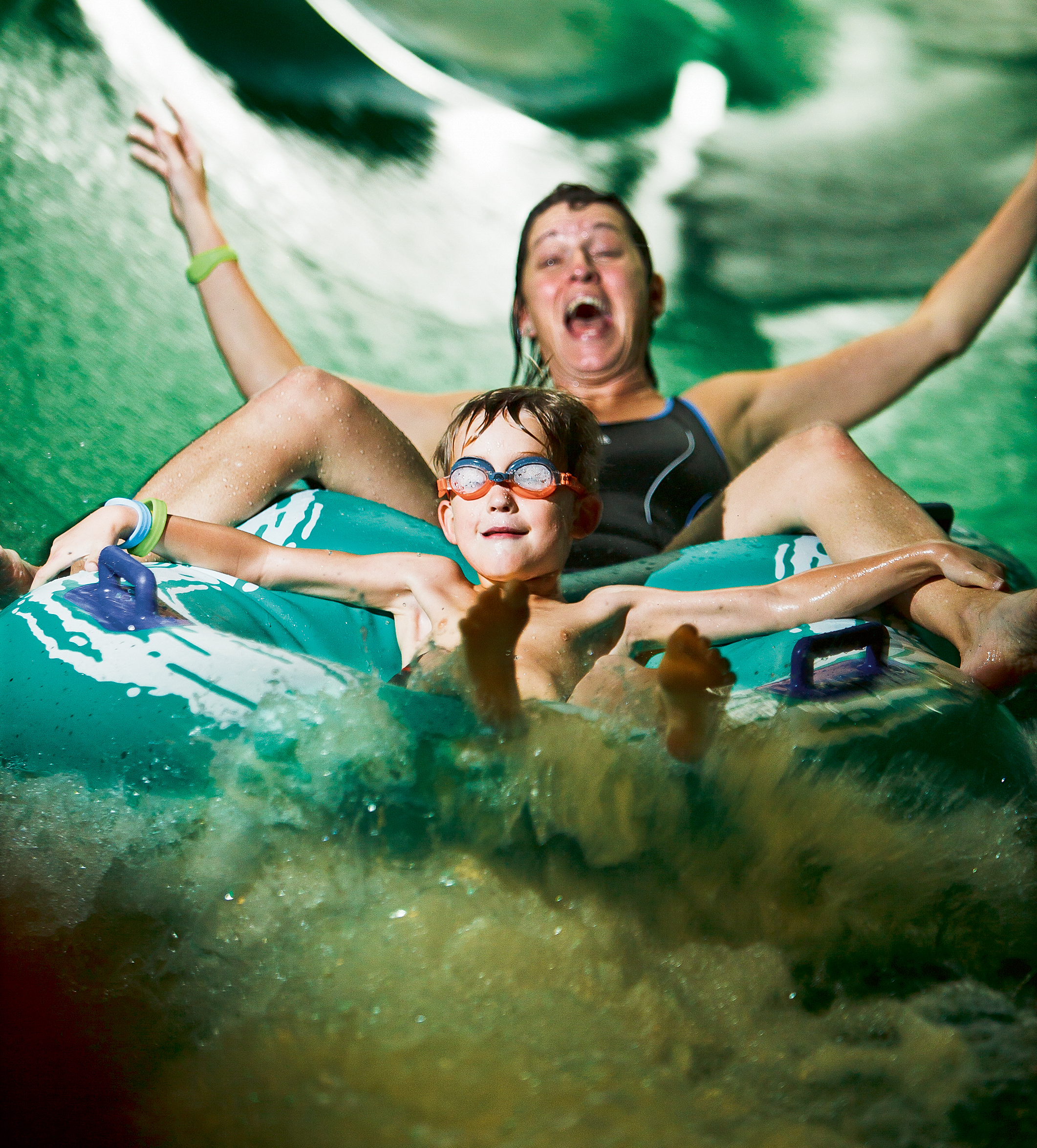 Två personer åker på en grön ring i en grön vattenrutschbana. De båda sträcker ut armarna och ser väldigt glada ut. Det stänker vatten överallt. 