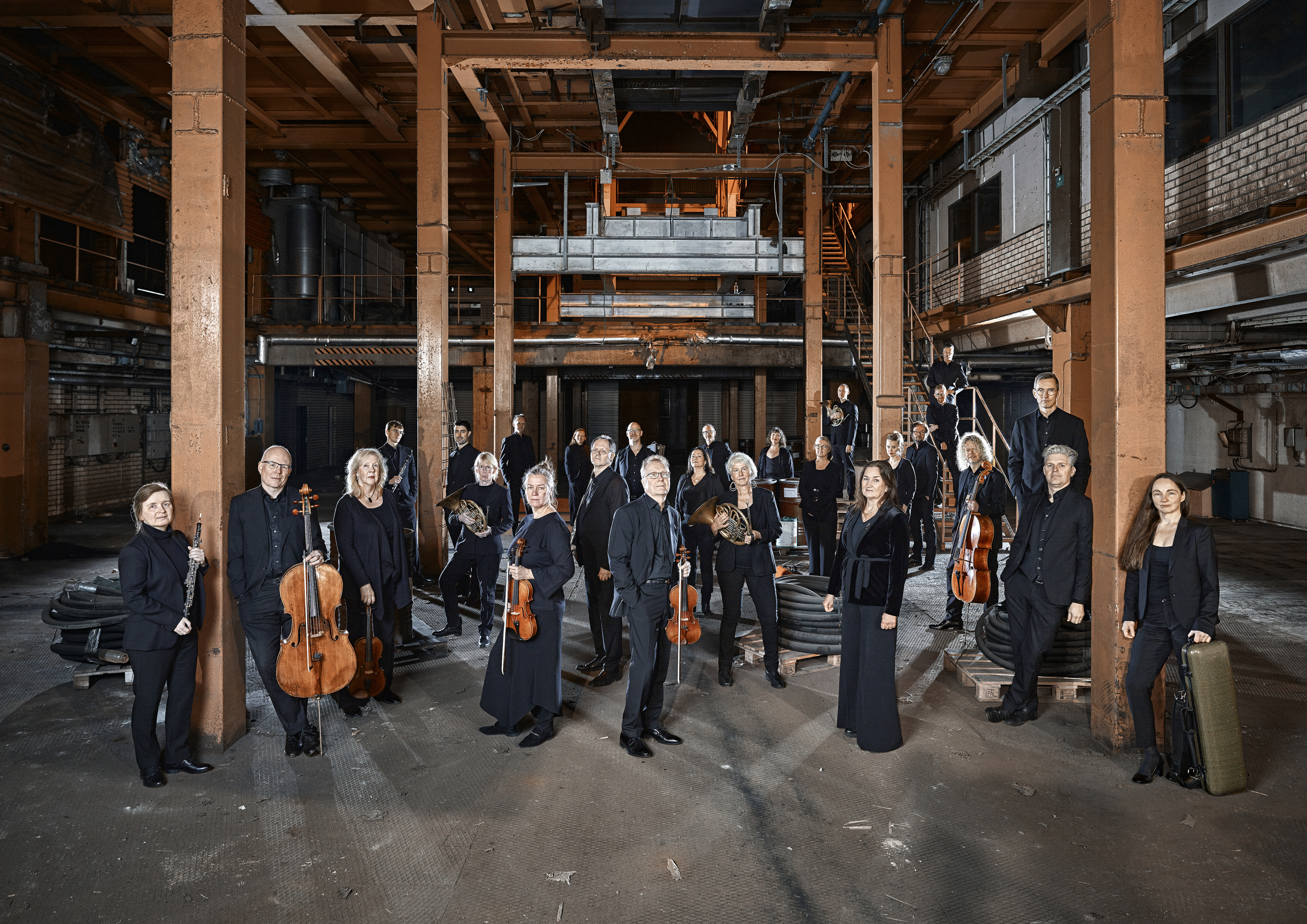 En bild på alla personer i Nordiska Kammarorkestern. Alla är iklädda svarta kläder och står med sina olika instrument i en stor öppen byggnad.