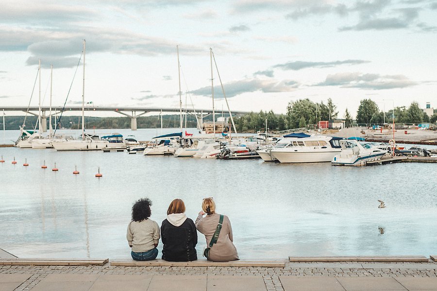 Tre personer sitter på en träbänk vid Sundsvalls småbåtshamn. Havet är spegelblankt och på himlen svävar några vita moln. I hamnen är det fullt med både vita segelbåtar och motorbåtar.