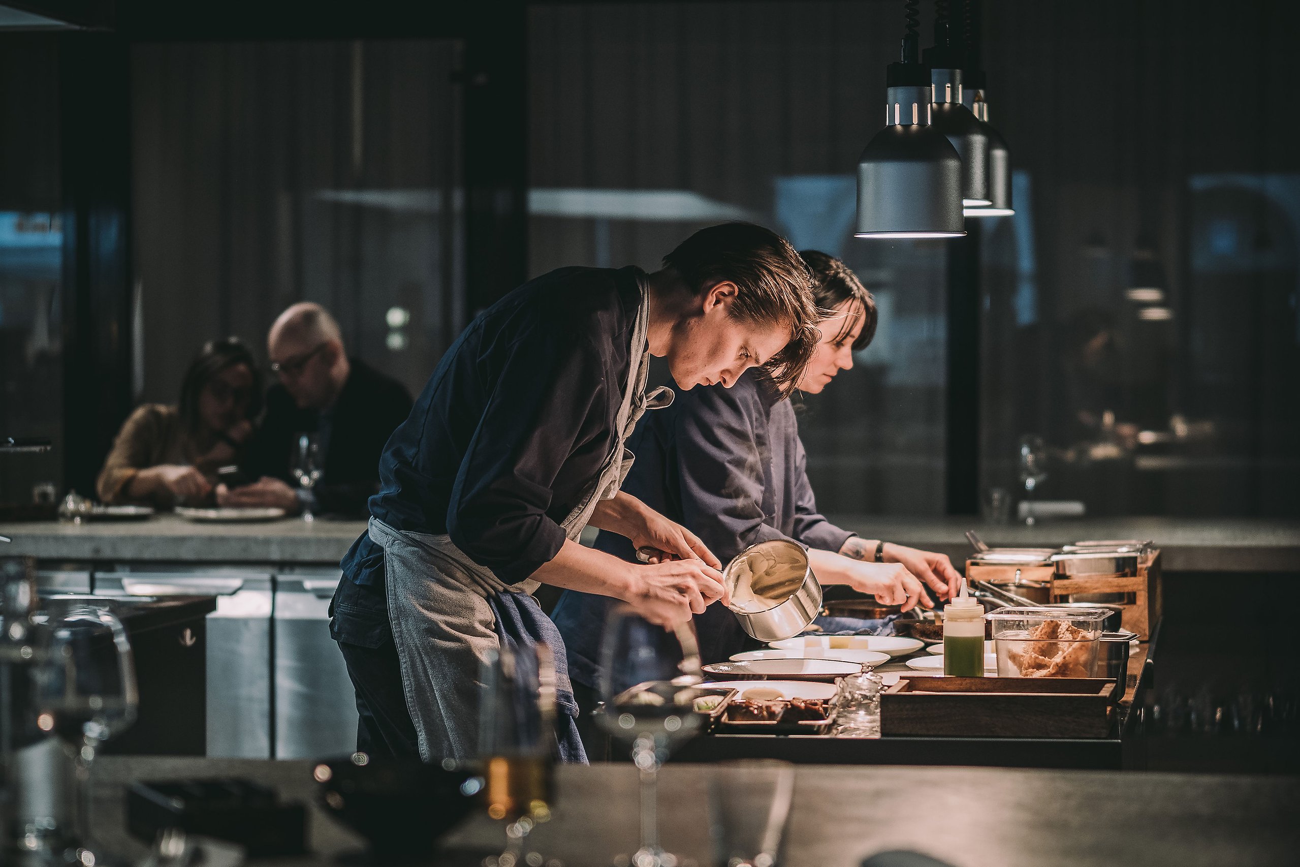 Foto av två personer som lagar mat i ett restaurangkök. Kockarna lägger upp mat väldigt noggrant. I bakgrunden sitter några människor och väntar på att bli serverade.
