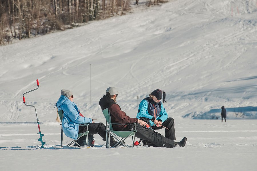 Fotografi av tre personer som sitter i campingstolar på isen och pimplar. Isen är täckt av snö. Personerna har på sig täckbyxor, jackor och mössor. Bakom dem står en isborr. 